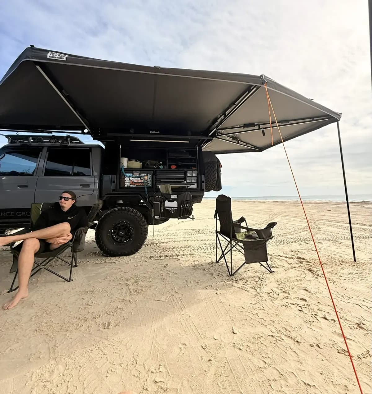 Sam Drew relaxing in a camp chair on the beach under the awning of his dark grey 79 Series, canopy open