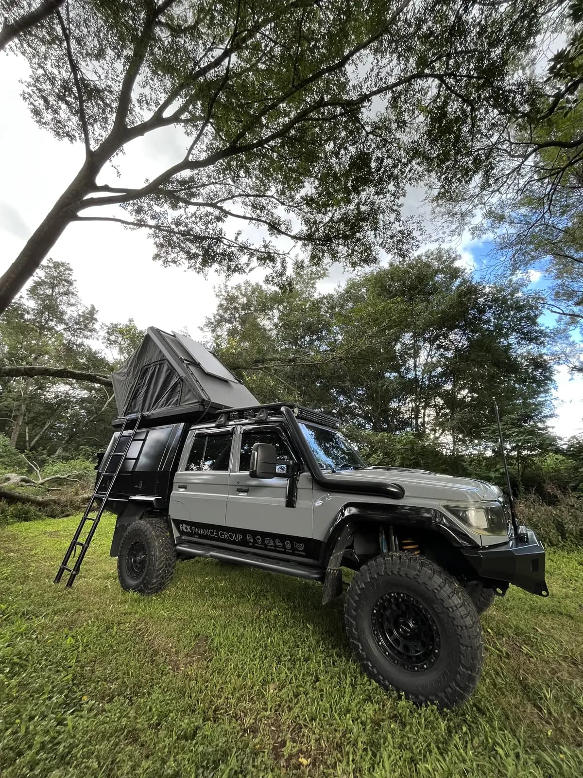 A dark grey 79 Series with its roof-top tent open and ladder down, set up for camp on a grassy riverbank under gum trees