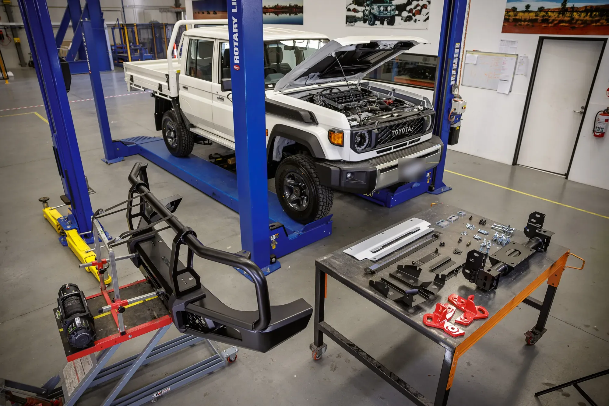 A white 79 Series dual-cab tray on a workshop hoist, bonnet up, with a new bullbar, winch and fitting parts laid out on the floor