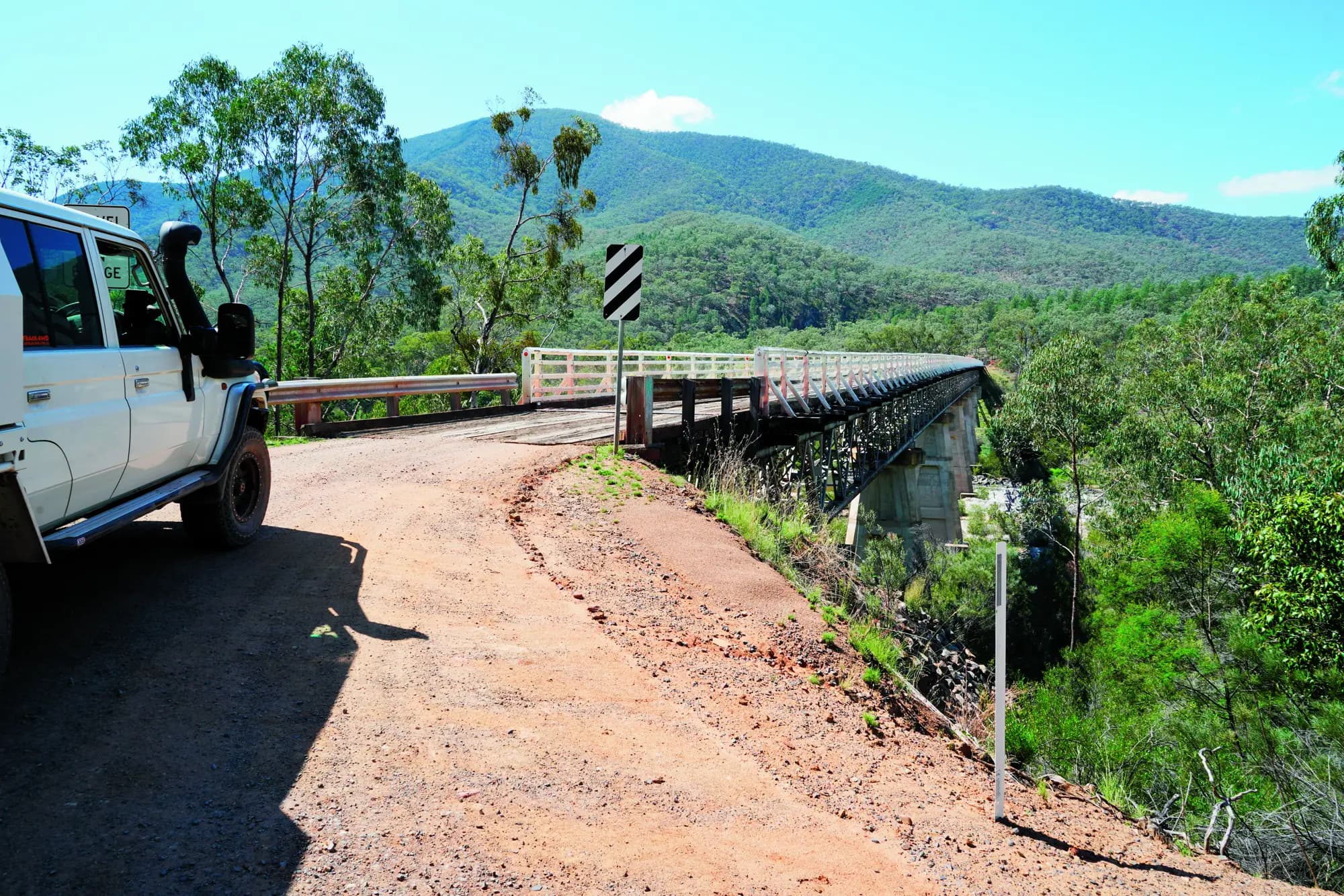 A white 4x4 with a snorkel pulled up at the start of an old timber bridge on a gravel road, forested ranges behind