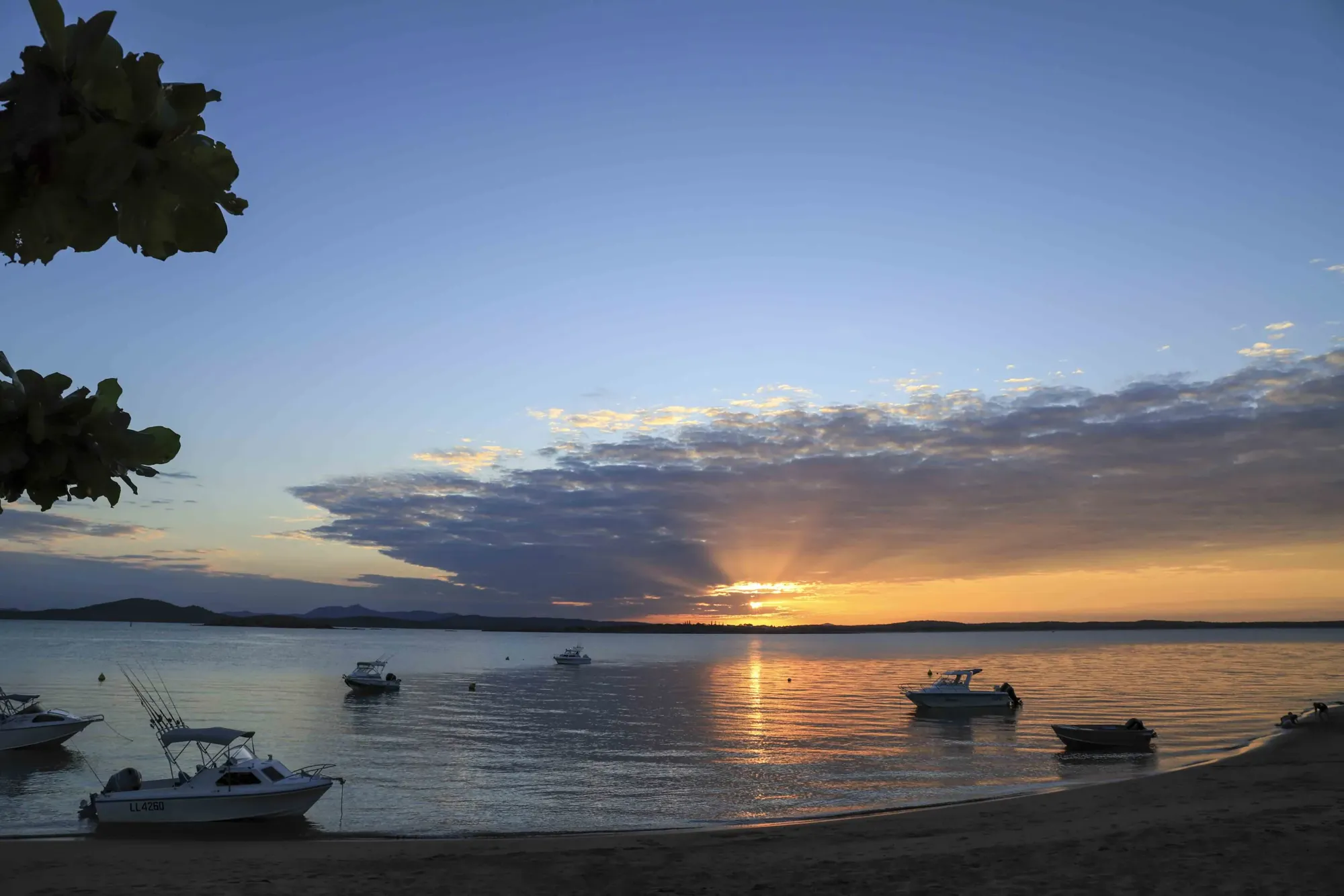 Fishing boats moored in a calm Queensland bay at sunset, sun breaking under the cloud on the horizon