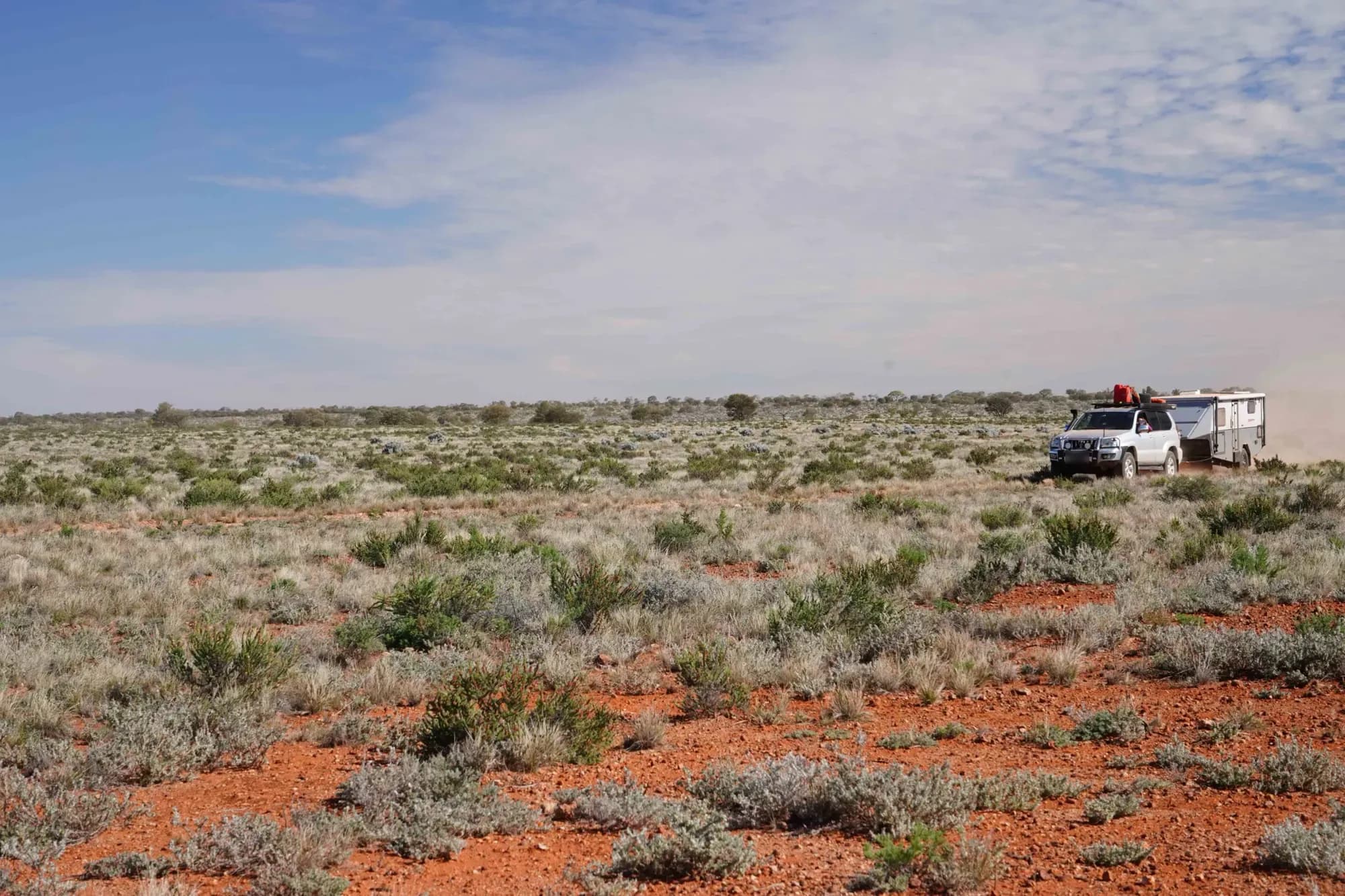 A 4x4 towing a caravan across open saltbush plains on red dirt under a wide blue sky