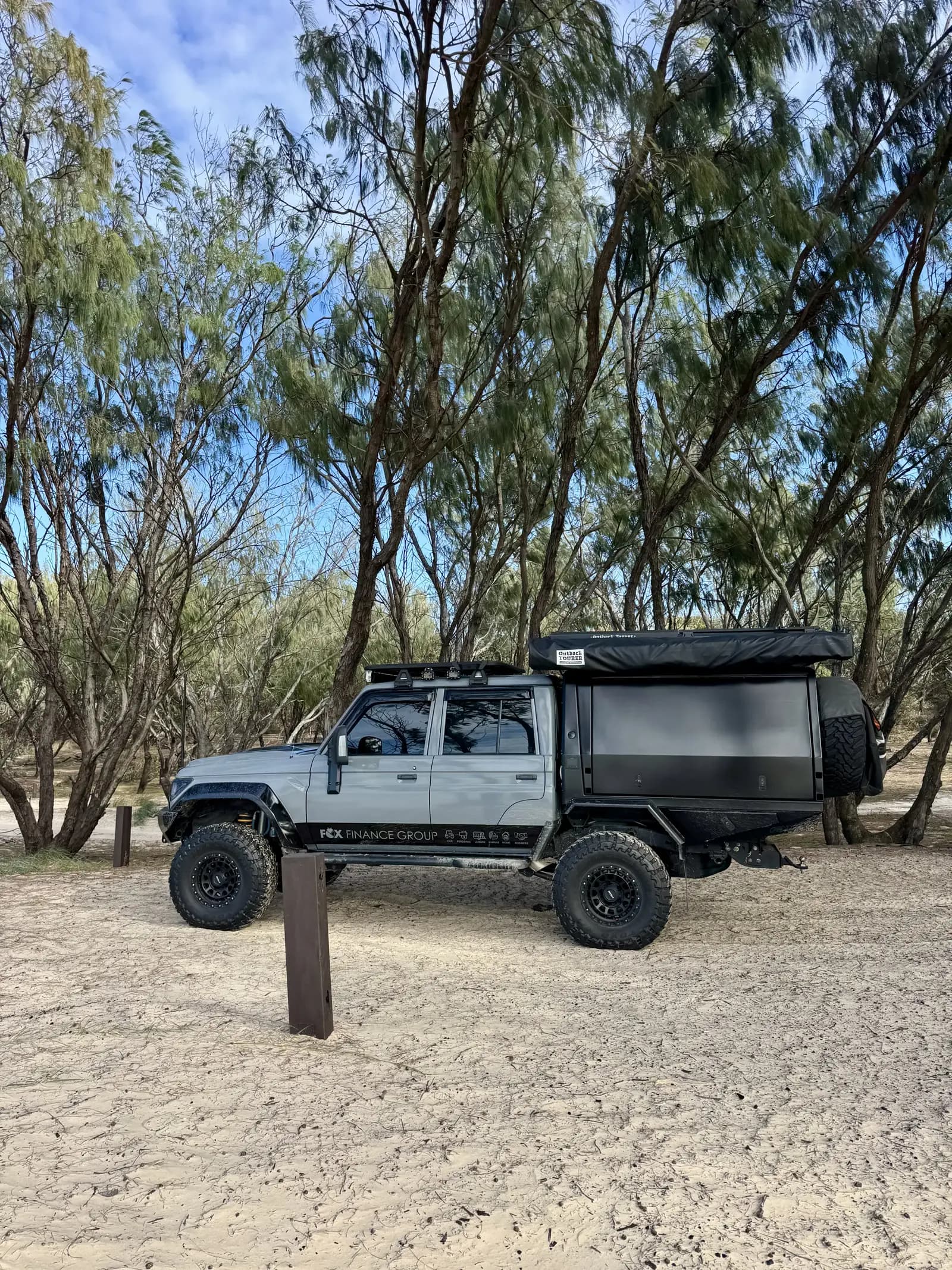 A dark grey 79 Series dual-cab with a black tray canopy and roof rack parked on beach sand under casuarina trees