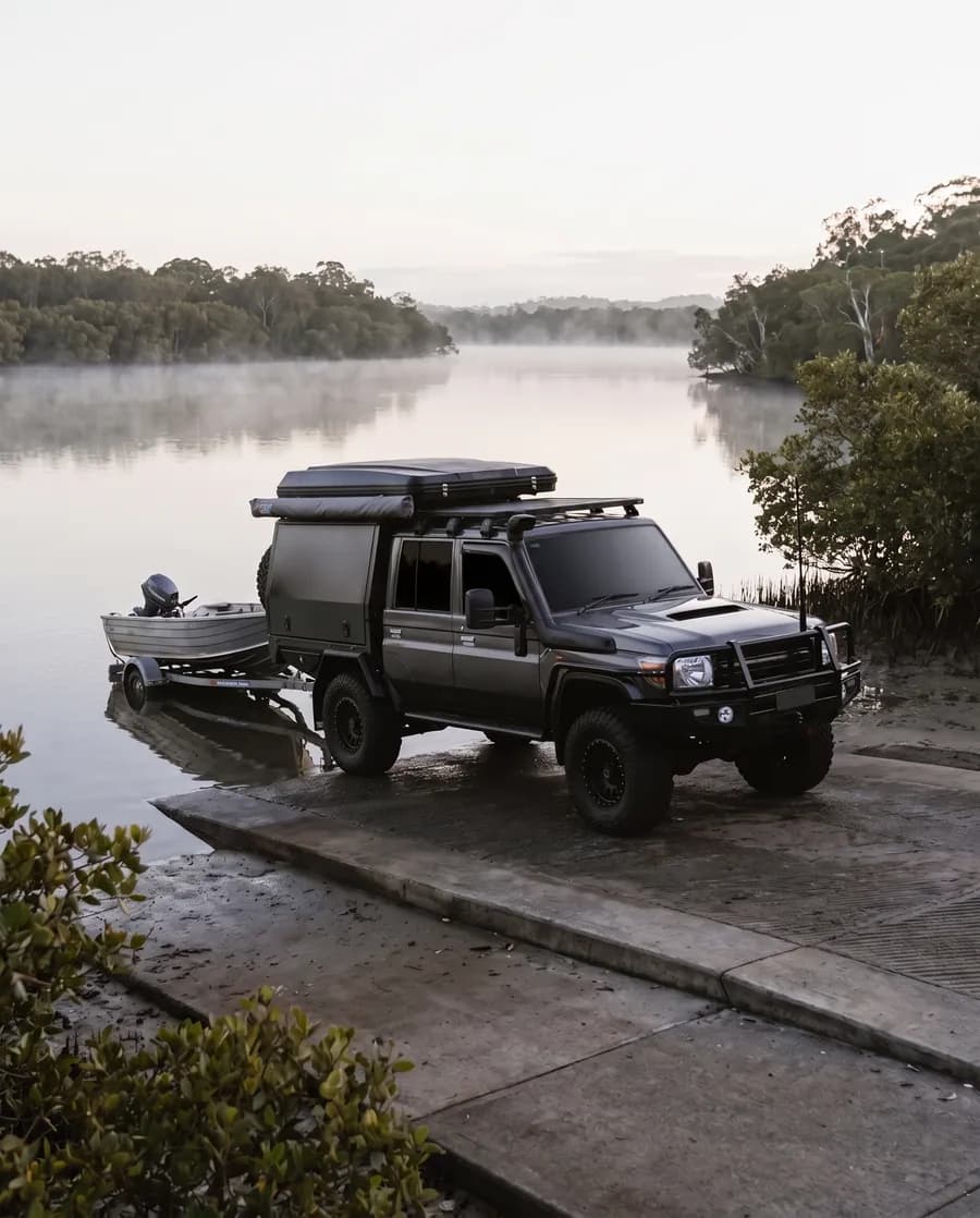 A dark grey dual-cab 79 Series with a black canopy and roof-top tent reversing a trailer with an aluminium tinny down a concrete estuary boat ramp at dawn, mangroves behind