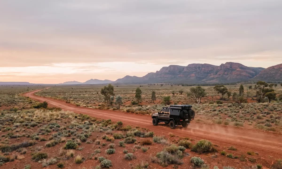 A dark grey dual-cab 79 Series with a black canopy and roof-top tent small on a long red dirt road crossing a saltbush plain with layered ranges behind
