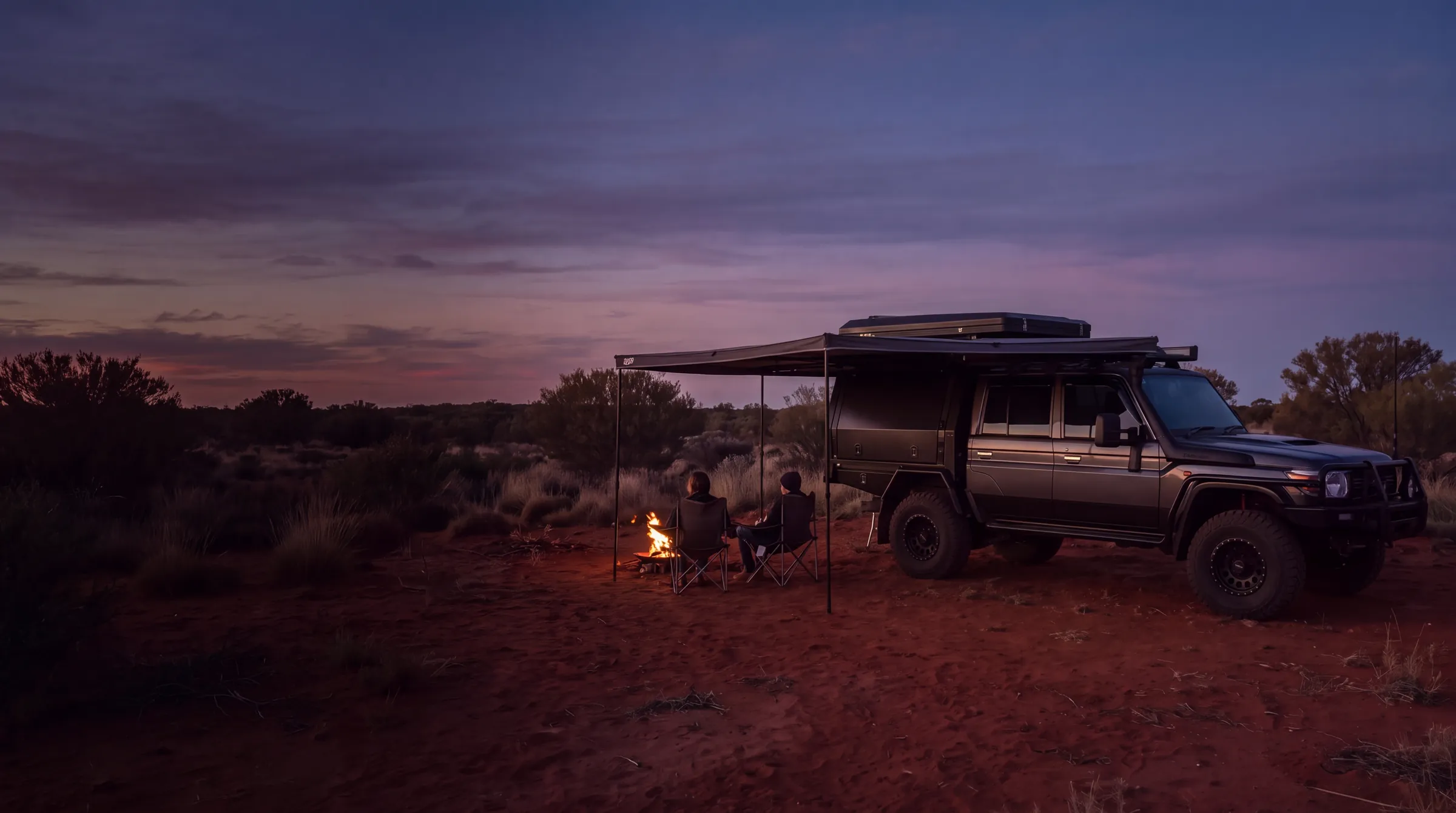 A dark grey dual-cab 79 Series with a black canopy and roof-top tent, its awning out at a red dirt outback camp at dusk, two people at a small campfire