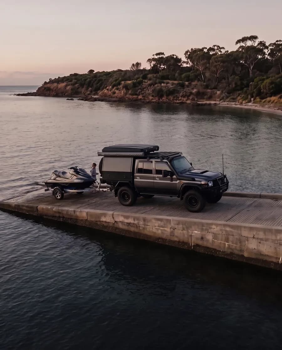 A jet ski on a trailer behind a dark grey dual-cab 79 Series with a black canopy and roof-top tent at the top of a concrete boat ramp on a calm bay at sunset