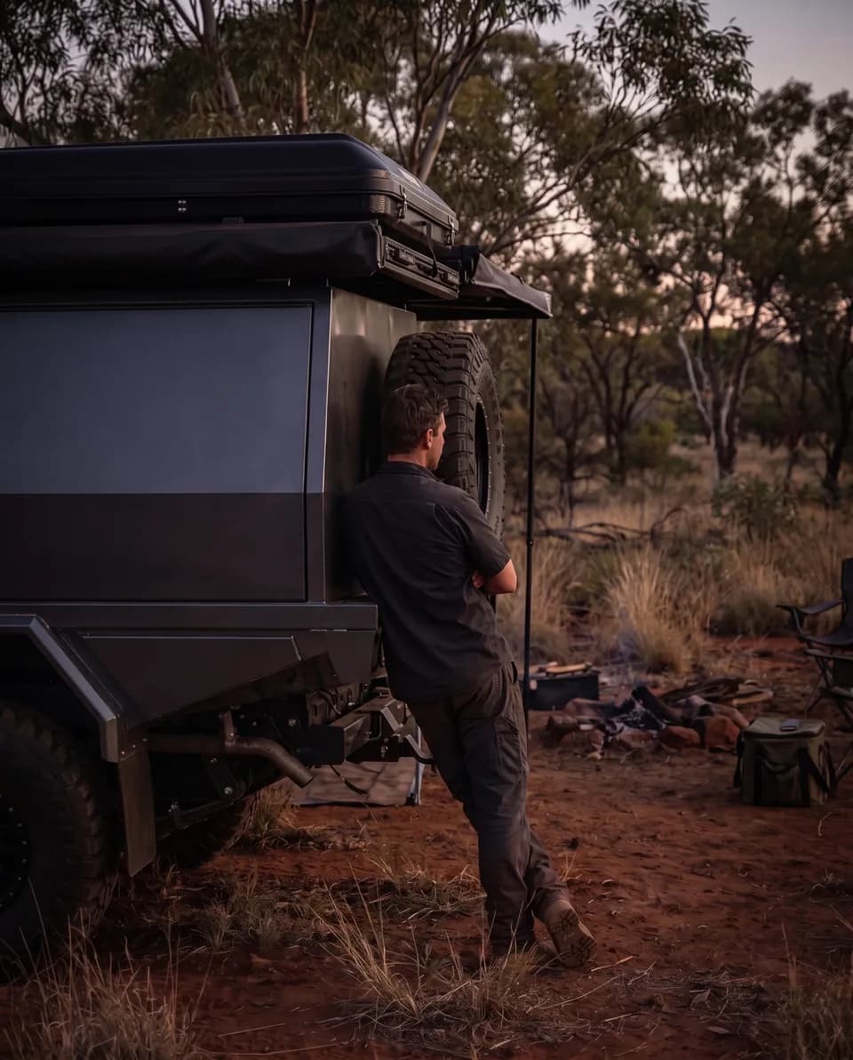 A person in a work shirt leaning on the tailgate of a dark grey dual-cab 79 Series with a black canopy and roof-top tent at a dusk bush camp, looking out at the bush