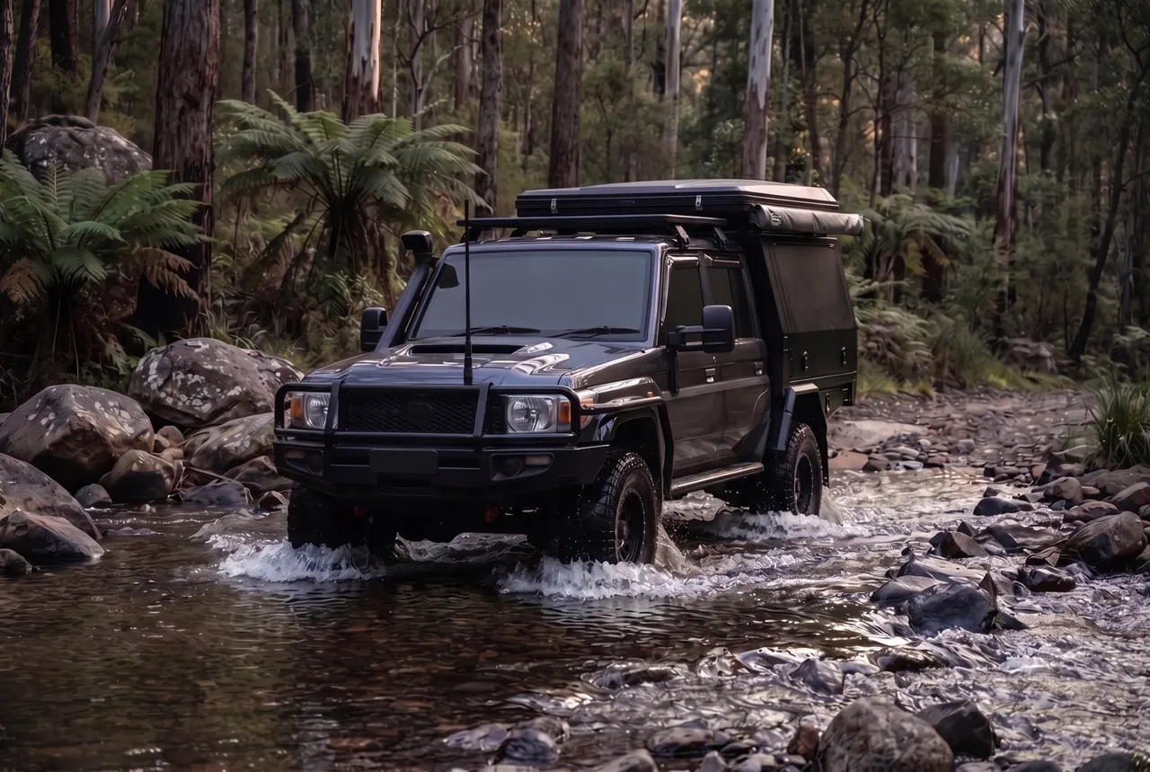 A dark grey dual-cab 79 Series with a black canopy and roof-top tent crossing a shallow rocky creek on a high country bush track, water pushing off the tyres