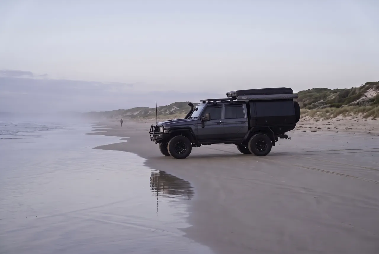 A dark grey dual-cab 79 Series with a black canopy and roof-top tent driving along the wet tide line of a hard-sand beach at dawn, one walker in the distance